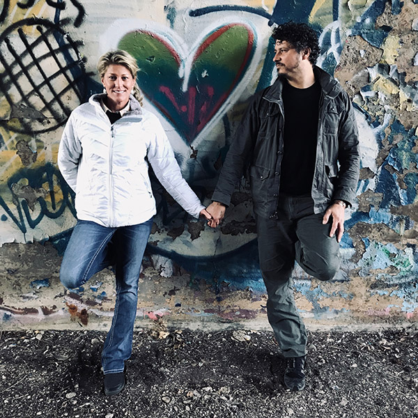 Man and woman stand in front of heart grafitti while holding hands