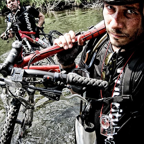 Two men carrying mountain bikes through a stream