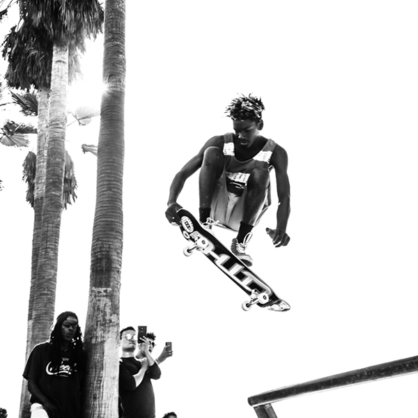Skateboarder jumps railing while spectators look on beneath palm trees